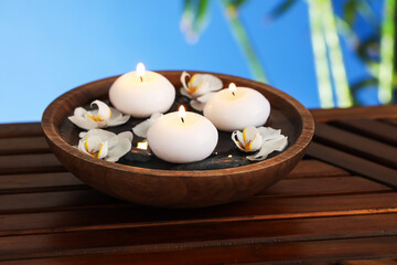 Burning candles and orchid flowers in bowl of water on wooden table against blurred blue...
