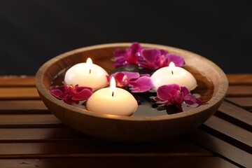 Burning candles and orchid flowers in bowl of water on wooden table against black background,...