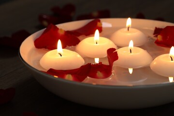 Burning candles and petals in bowl of water on wooden table, closeup