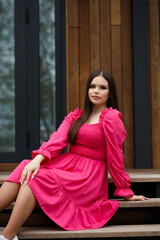 A woman sits on the steps of a wooden veranda outside a modern home. She wears a bright pink dress...
