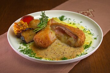 Crispy cordon bleu cutlet filled with ham and melted cheese, served with mustard sauce and grilled vegetables on a white plate.
