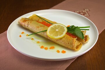 Delicate crepes filled with red caviar, garnished with lemon slice, herbs, and olive oil drops on a white plate.