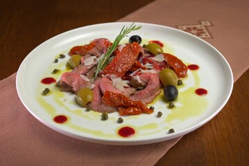Beef carpaccio with sun-dried tomatoes, olives, capers, parmesan shavings, and rosemary, drizzled with olive oil and berry reduction on a white plate.