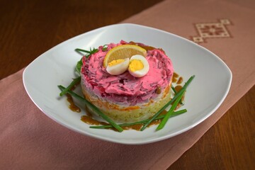 Layered herring salad with beetroot, carrots, potatoes, and mayonnaise, topped with quail egg and lemon slice, served on a white plate.
