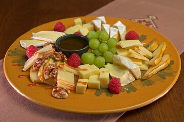 Cheese platter with assorted cheeses, fresh grapes, raspberries, pear slices, walnuts, and honey dip, elegantly served on a decorative plate.