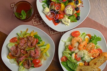 Top view of three fresh salads with grilled beef, shrimp, and Greek-style ingredients, served on white plates with a refreshing drink in a glass on a traditional embroidered napkin.