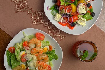 Top view of shrimp Caesar salad and Greek salad with iced tea on embroidered brown linen, featuring colorful fresh ingredients and rustic presentation.