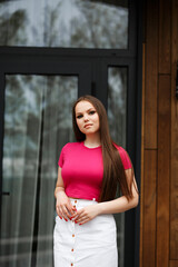 A woman standing on a wooden veranda in front of a modern house. She is wearing a bright pink...