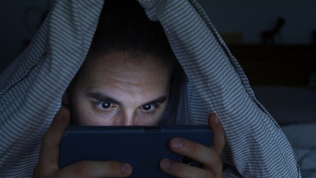 Smartphone and social network addiction concept. Young male using smartphone in bed with blanket over head