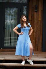 A woman stands on a veranda near a wooden wall of a building. She is wearing a blue dress with...