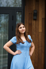 A woman stands on a veranda near a wooden wall of a building. She is wearing a blue dress with...