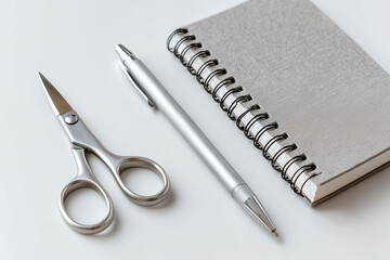 A pair of silver scissors, a sleek silver pen, and a gray spiral notebook are neatly arranged on a white surface, perfect for creative tasks.