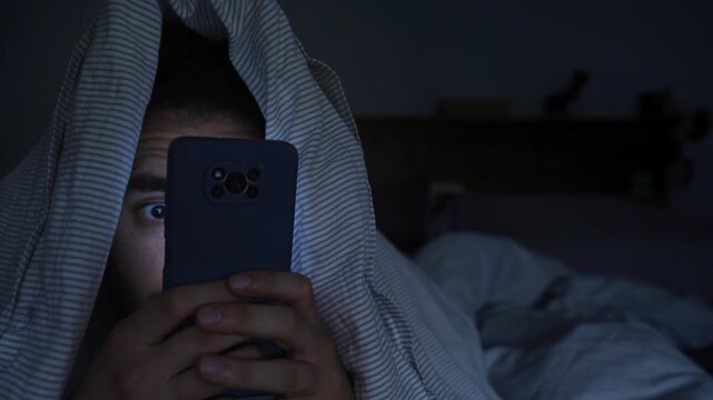 Smartphone and social network addiction concept. Young male using smartphone in bed with blanket over head