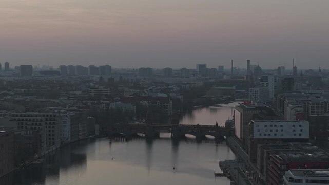 Approaching aerial toward the Oberbaum Bridge in Berlin at vivid pre dusk sunset with a subway train crossing clear sky warm water reflections moving traffic and the waterfront architecture of Kreuzbe