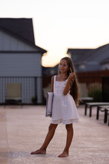 young girl in a white dress holding a notebook, standing outdoors barefoot in the evening sunlight.