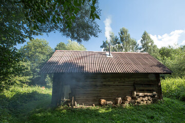 Fototapeta premium A bathhouse located in a forest clearing. The walls are made of logs, and the roof is covered with old slate. Smoke comes out of the chimney sticking out of the roof. 