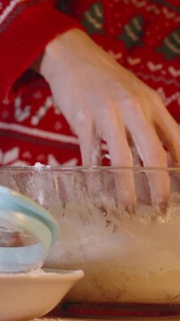 Vertical video. Woman in Red Holiday Sweater Mixing Dough with Hands in Glass Bowl on Wooden Table in Cozy Kitchen with Soft Lighting