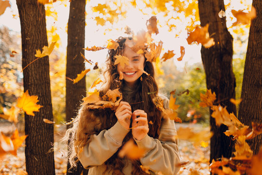 Smiling positive young woman throws up the yellow autumn leaves in the forest at sunset