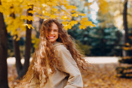 Smiling young woman with curly hairstyle enjoys the autumn weather in the forest with the yellow leaves at sunset