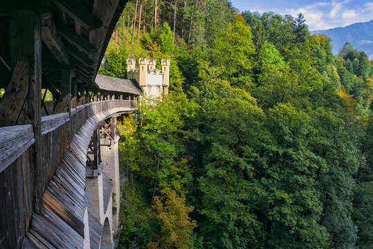 St. Georgenberg, Austria: A view of a rustic, covered wooden walkway or bridge leading into a lush green forest, with a small stone tower visible in the background.