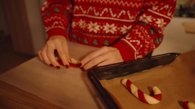 Woman Carefully Adjusting Candy Cane Cookie Shape on Parchment Paper Near Baking Tray with Red and White Dough