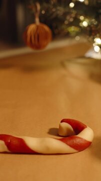 Vertical video. Woman Shaping Candy Cane Cookie Curve with Fingers Beside Decorated Christmas Tree and Red Santa Hat