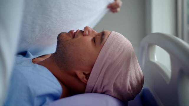 close up side view of dead patient man with cancer in headscarf lying on bed having covering with white sheet by doctor on dead male at hospital ward,
illness and the end of life concept.