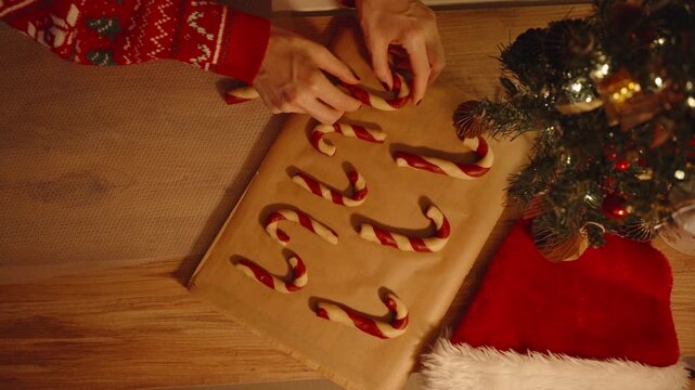 Woman Aligning Candy Cane Cookies in Rows from Overhead View with Christmas Tree and Santa Hat Beside