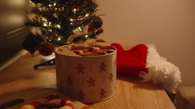 Large Tin with Candy Cane Cookies on Wooden Table beside Santa Hat and Christmas Tree with Lights