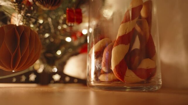 Low Angle Close-Up of Glass Jar Filled with Candy Cane Cookies by Christmas Tree in Warm Ambient Light