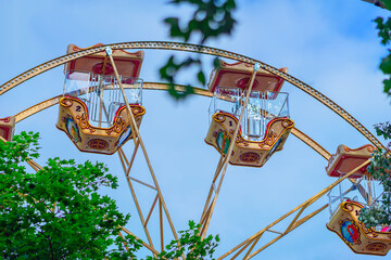 Ferris Wheel Cabins. Close-up view of ornate Ferris wheel cabins framed by green tree leaves against blue sky. Concept of amusement park ride, summer leisure activity, attractions