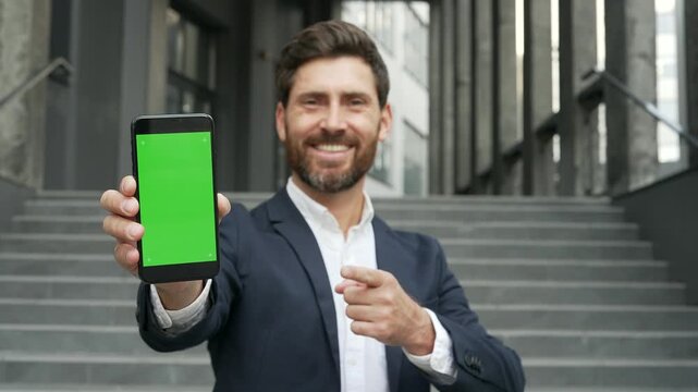 Happy businessman showing mobile phone with green screen standing on street near office building. Color key, vertical template layout for advertising. Smiling male looking at camera holding smartphone