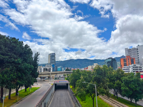 Vibrant panoramic view of Bogot&aacute;, Colombia, showcasing Calle 100 with modern buildings, green trees, and mountains in the background under a partly cloudy sky