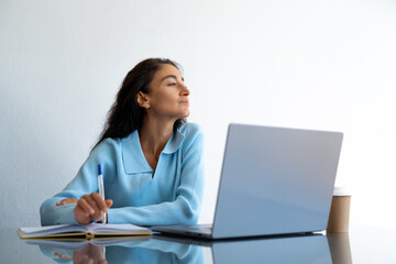Middle-aged woman with long dark hair in a light blue sweater holding a pen and looking thoughtfully to the side while sitting at a glass desk with laptop, notebook, and coffee cup in bright office