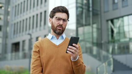 Shocked worried businessman reading bad news on mobile phone while standing on the street near an office building. Frustrated stressed entrepreneur worker has a bad mood, looking at negative message - Powered by Adobe