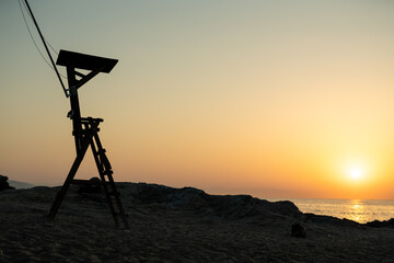 Wooden lifeguard tower silhouette against a golden sunrise over the ocean, with rocky coastline and sandy beach in the foreground