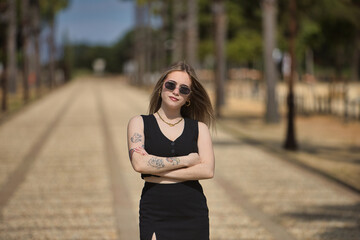 Young, beautiful, blonde woman, with black top and skirt and sunglasses, with her arms crossed,...