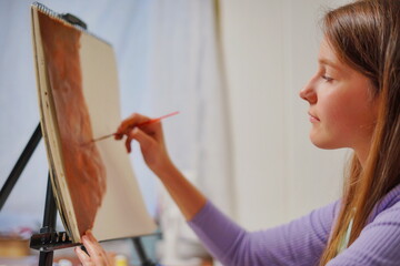 Close-up of a little girl painting in a sketchbook at a desk with an easel in art studio