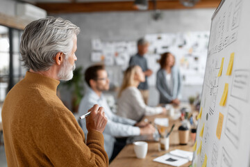 A distinguished senior male colleague leads a brainstorming session, presenting ideas on a whiteboard to a diverse team of professionals in a modern, light-filled office environment.

