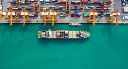 Aerial view of a loaded cargo ship leaving a port, with cranes and shipping containers visible.
