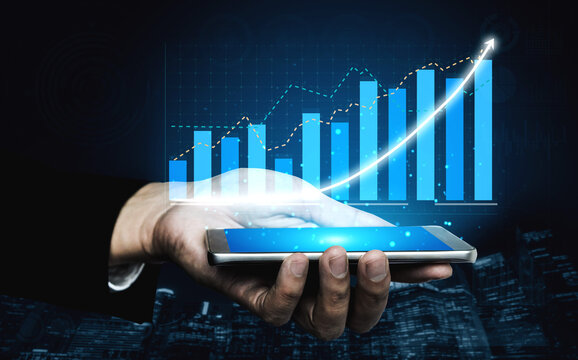 A close-up view of a hand holding a smartphone showing a financial growth graph with blue bars and a trend line. The background features a dark cityscape. Impute
