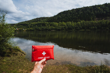A hand is gently holding a bright red first aid kit while positioned by a serene and tranquil lake surrounded by lush nature, showcasing the beauty of the landscape and inviting outdoor exploration