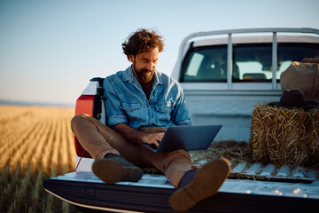 Smiling man using laptop while sitting in back of pick up truck in field. © Drazen