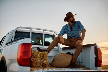 Smiling farmer sitting on hay bale in back of his truck at sunset.