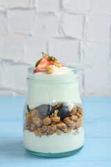 Tasty yogurt with granola and berries in glass jar on light blue wooden table, closeup