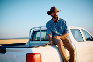 Pensive man sitting on his truck in farm field and looking away.