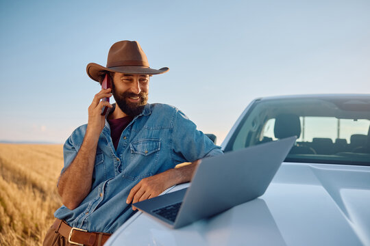 Happy farm worker talking on cell phone while working on laptop in field.