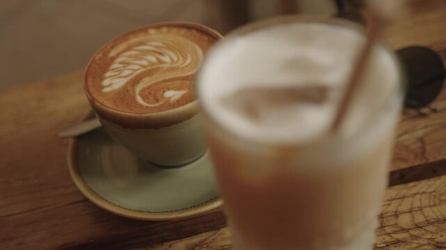 Cappuccino with latte art and blurred iced latte in the foreground on a rustic wooden caf&eacute; table