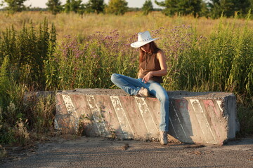 Beautiful young woman in jeans and hat sitting on a concrete block by the road