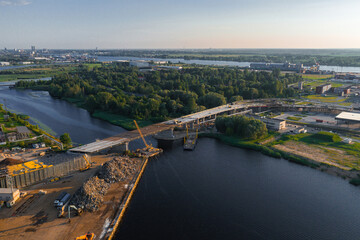 A partially completed bridge spans a river in Riga, Latvia, with cranes, construction materials,...